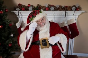 Santa sitting in front of a fireplace, next to a christmas tree